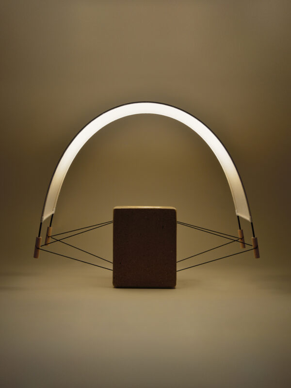 Vue de biais de la petite lampe à poser Écla de la Collection Aura créée par Maison Cédrat. Elle se compose d'un pied en brique et d'un abat jour en papier washi. La lampe est en finition terre crue cirée beige. allumée elle diffuse une lumière douce et dorée presque végétale dans une ambiance chaleureuse.