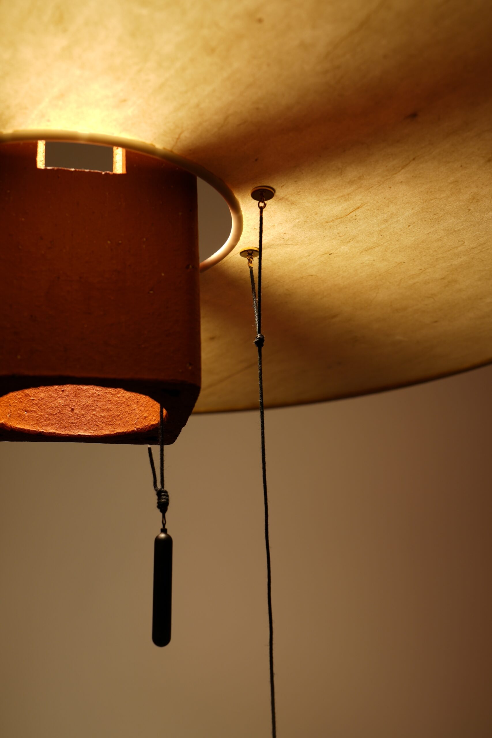 Image du détail de la lampe Nova allumée, on distingue la lumière qui se diffuse à travers une forme ajourée en terre rouge et traverse un anneau en papier washi. Les fribes du papier se révèlent dan un subtile jeu d'ombres et de lumière.