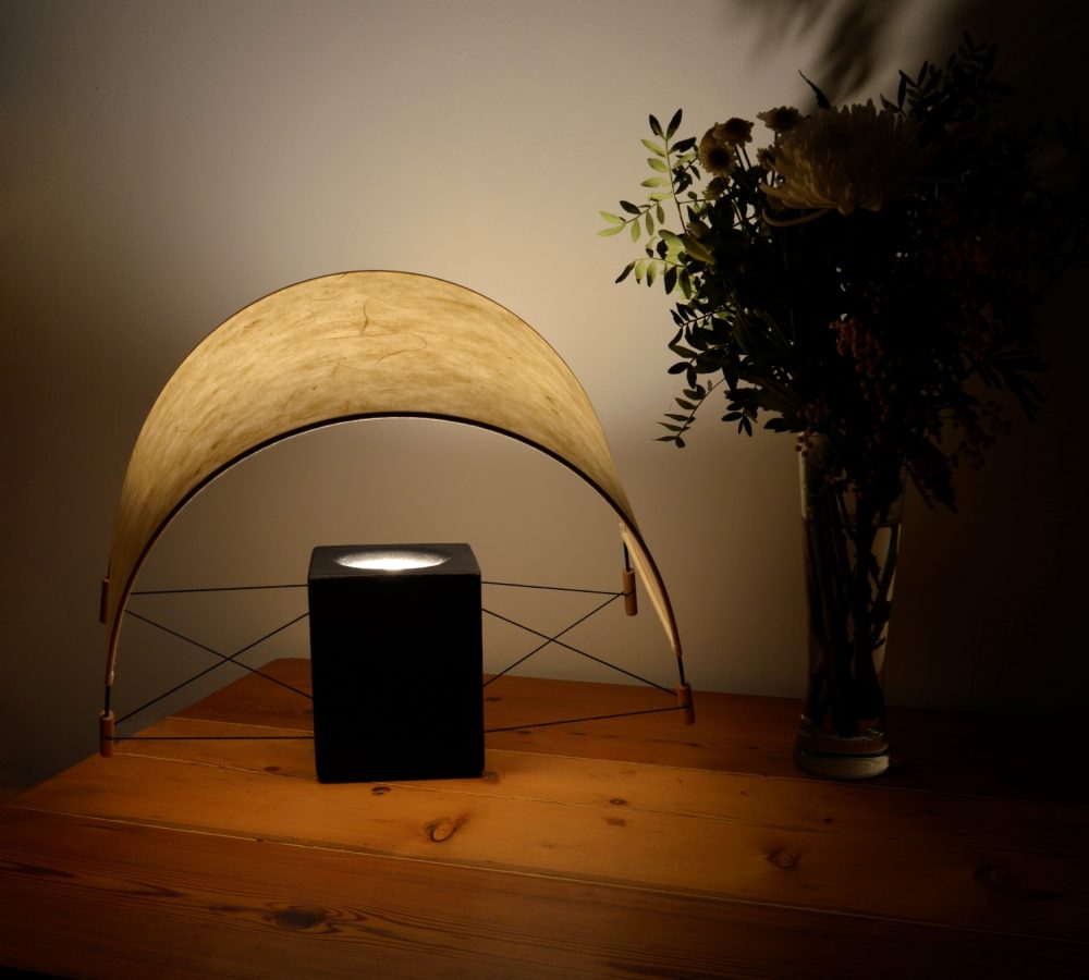 Lampe à poser Écla Noire allumée dans un chalet, avec son abat-jour en papier washi légèrement incliné vers l'avant.