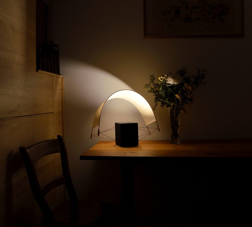Écla Noire allumée avec la voile orientée vers l'arrière posée sur la table d'un chalet en bois. La lampe diffuse une lumière intime et chaleureuse.