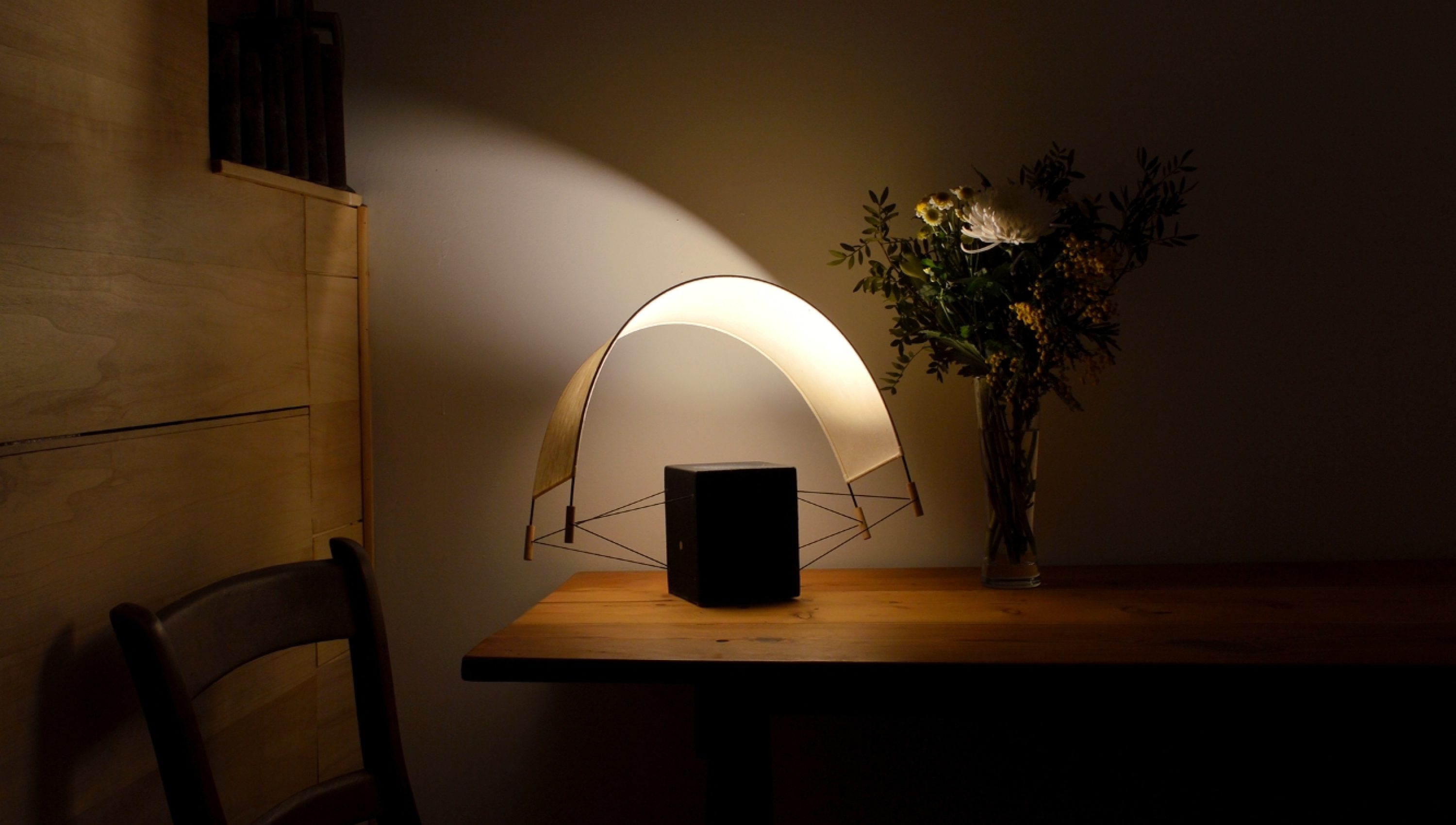 Écla Noire allumée avec la voile orientée vers l'arrière posée sur la table d'un chalet en bois. La lampe diffuse une lumière intime et chaleureuse.