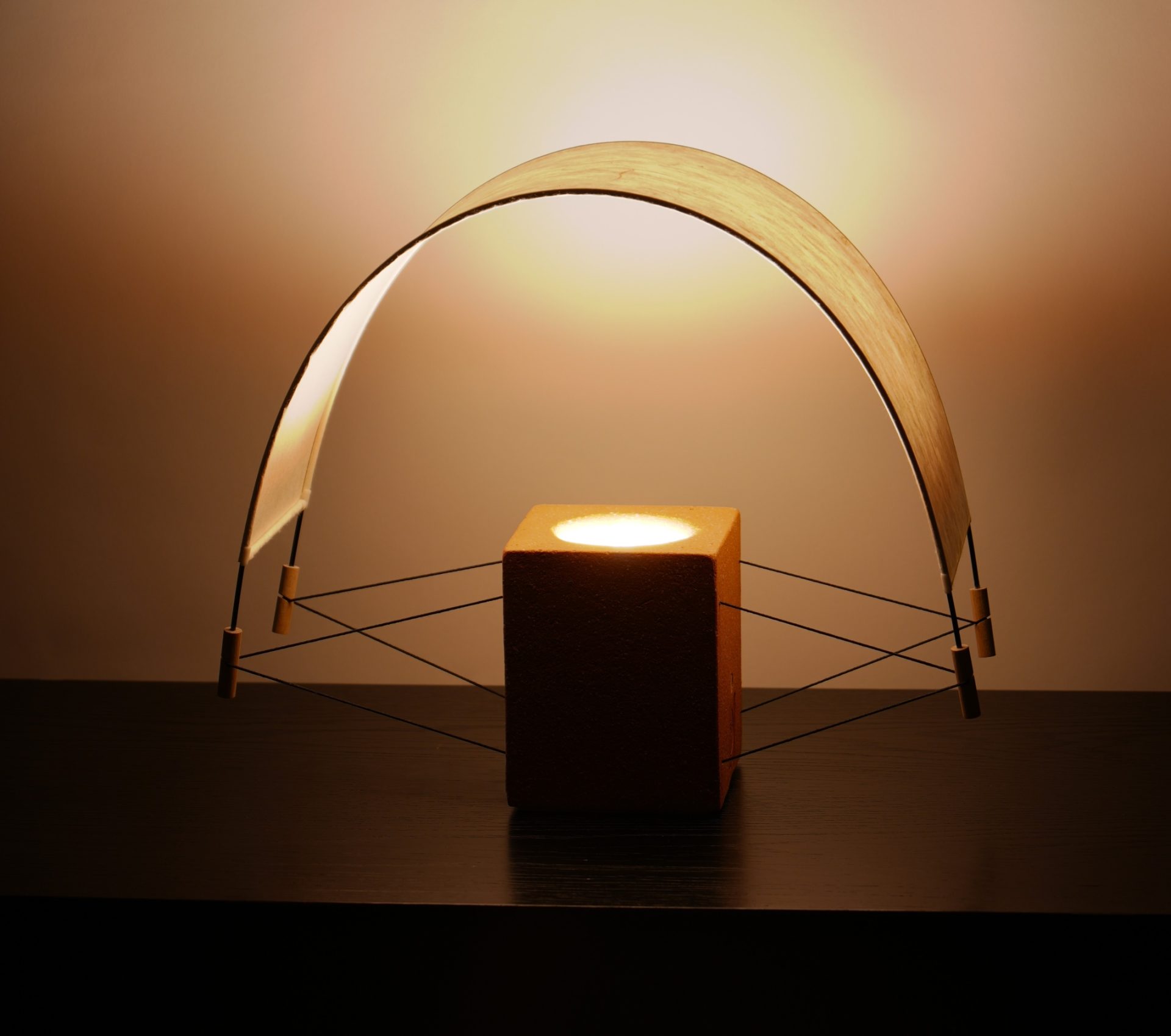 Photographie de la lampe à poser Écla vue de biais. En finition terre cuite rouge, on discerne une silhouette plus design entre le pied en terre noir et la voiles en papier japonaise mise en tension. La lumière jaune pâle est douce et se diffuse de manière chaleureuse.